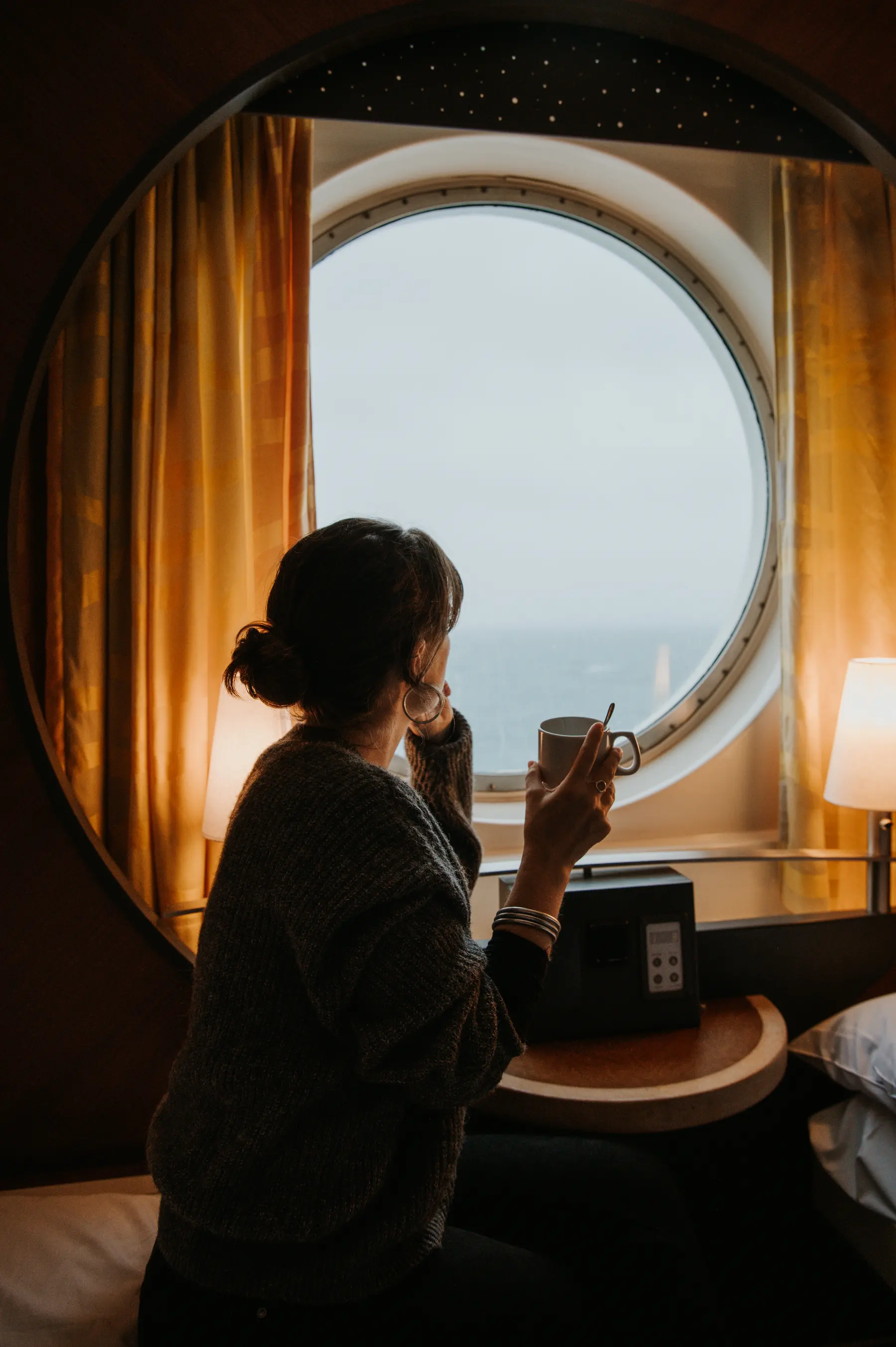 Une femme boit un café près du hublot d’une cabine.