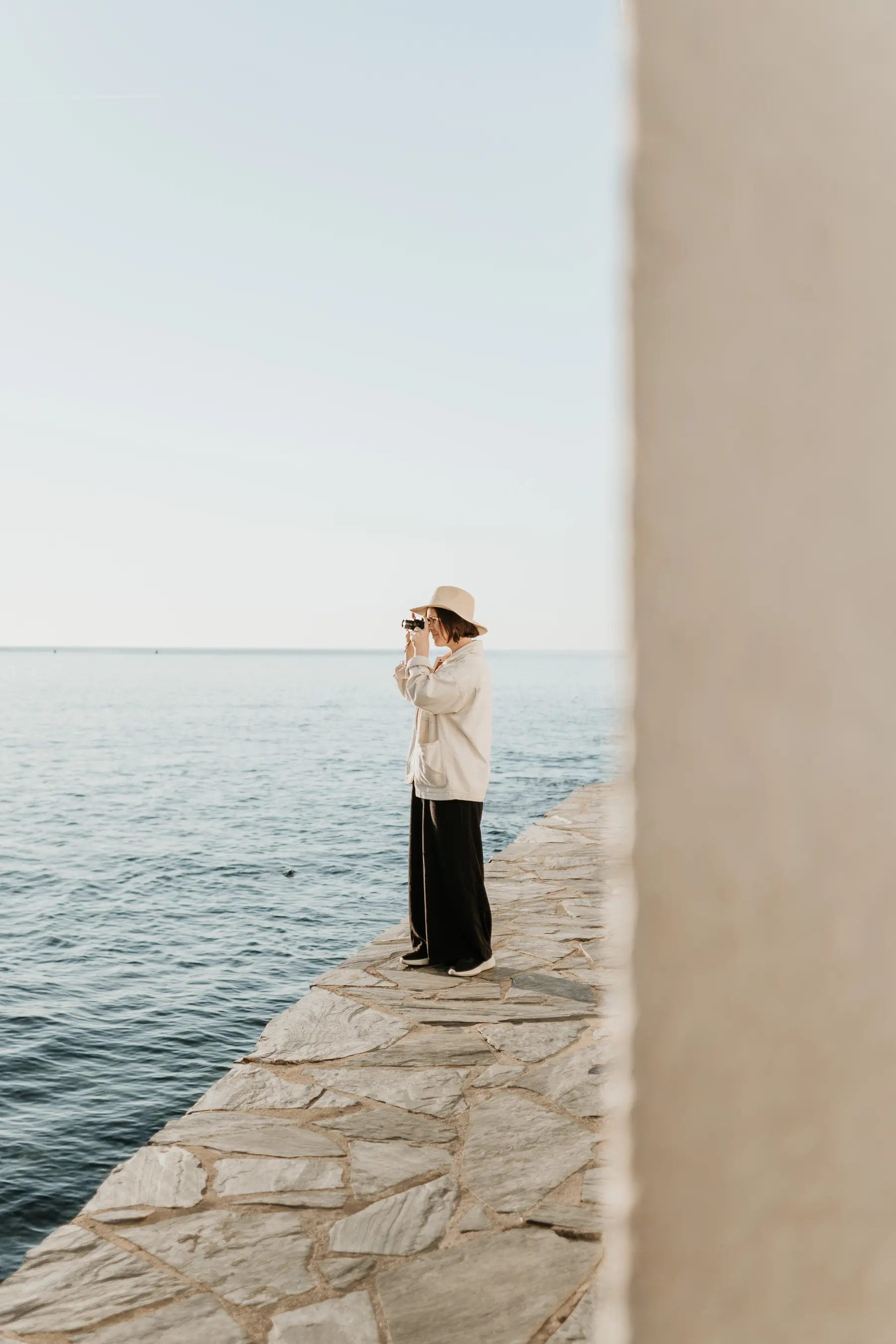 Une femme au chapeau attend sur un quai face à la mer.